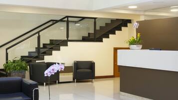A modern lobby with a reception desk, black leather sofas, a staircase, and a hanging light fixture.