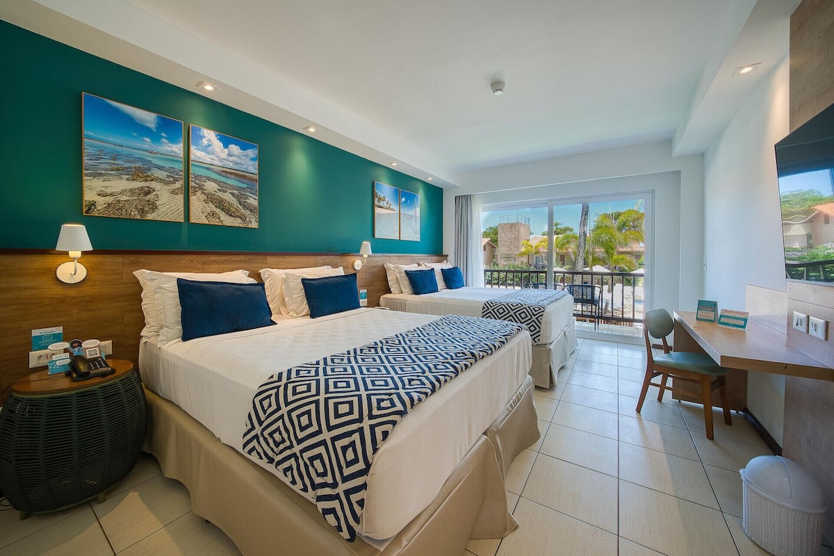 A hotel room with two beds, a desk, a chair, a balcony with a view, and artwork on the walls.