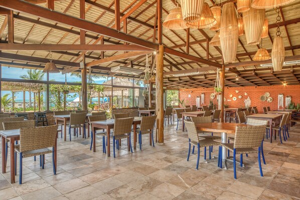 An indoor dining area with wooden tables and chairs, a high ceiling with exposed beams, and hanging light fixtures. - Salinas Maceió All Inclusive Resort (Maceió)