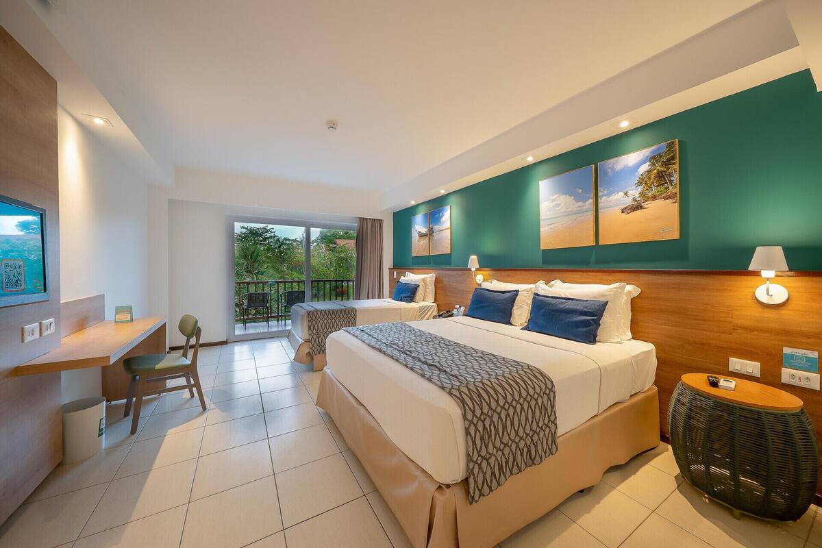 A hotel room with a large bed, a desk, a chair, a balcony with a view of greenery, and artwork on the walls.