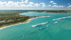 An aerial view of a coastal area with a beach, clear turquoise waters, and a distant town. - Salinas Maceió All Inclusive Resort (Maceió)
