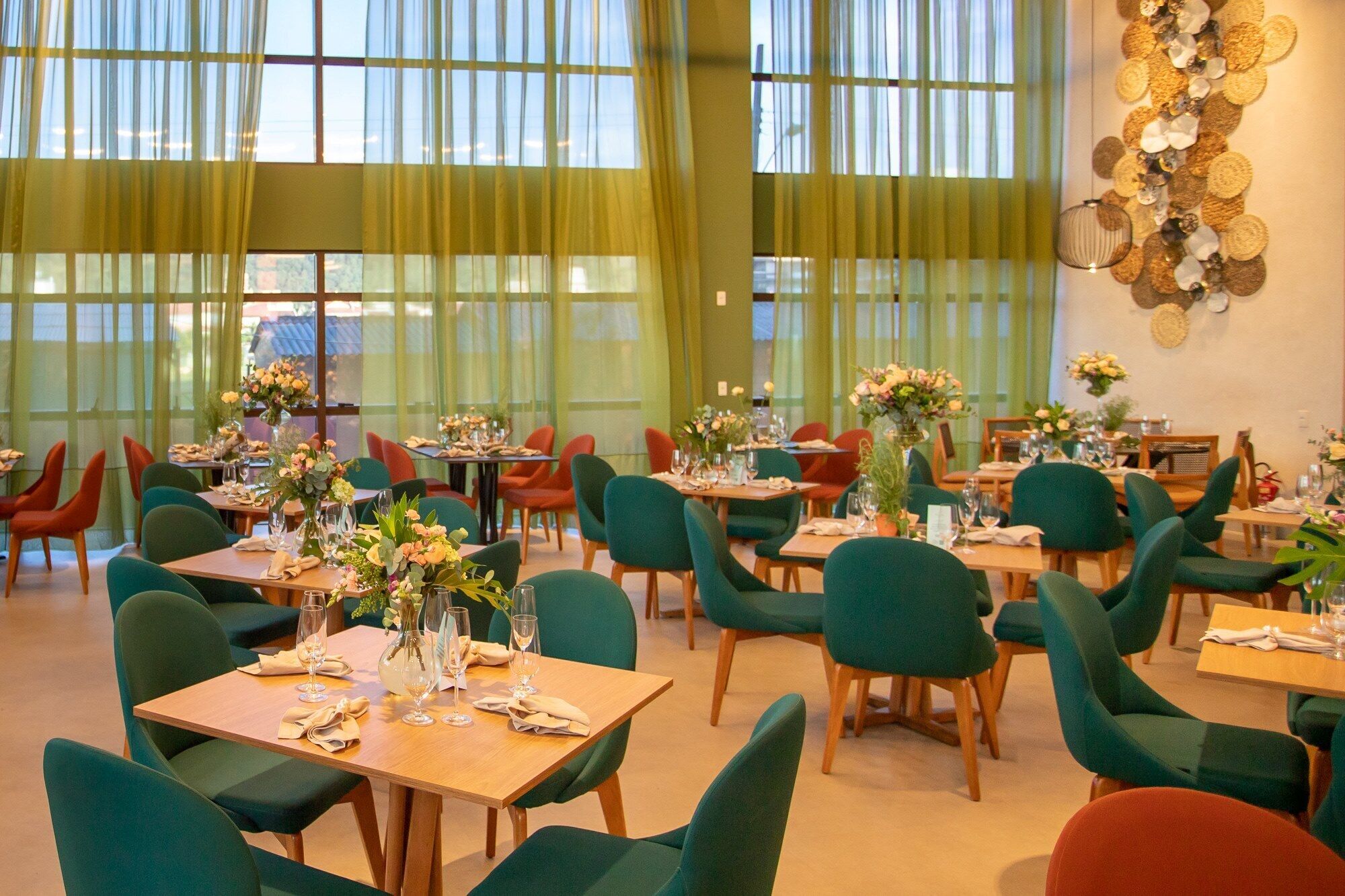 Een restaurant met meerdere tafels en stoelen, grote ramen met groene gordijnen en decoratieve muurkunst.