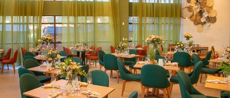 Een restaurant met meerdere tafels en stoelen, grote ramen met groene gordijnen en decoratieve muurkunst.