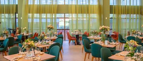 Un comedor amplio con mesas dispuestas para el servicio, ventanas grandes con cortinas translúcidas y lámparas colgantes circulares.