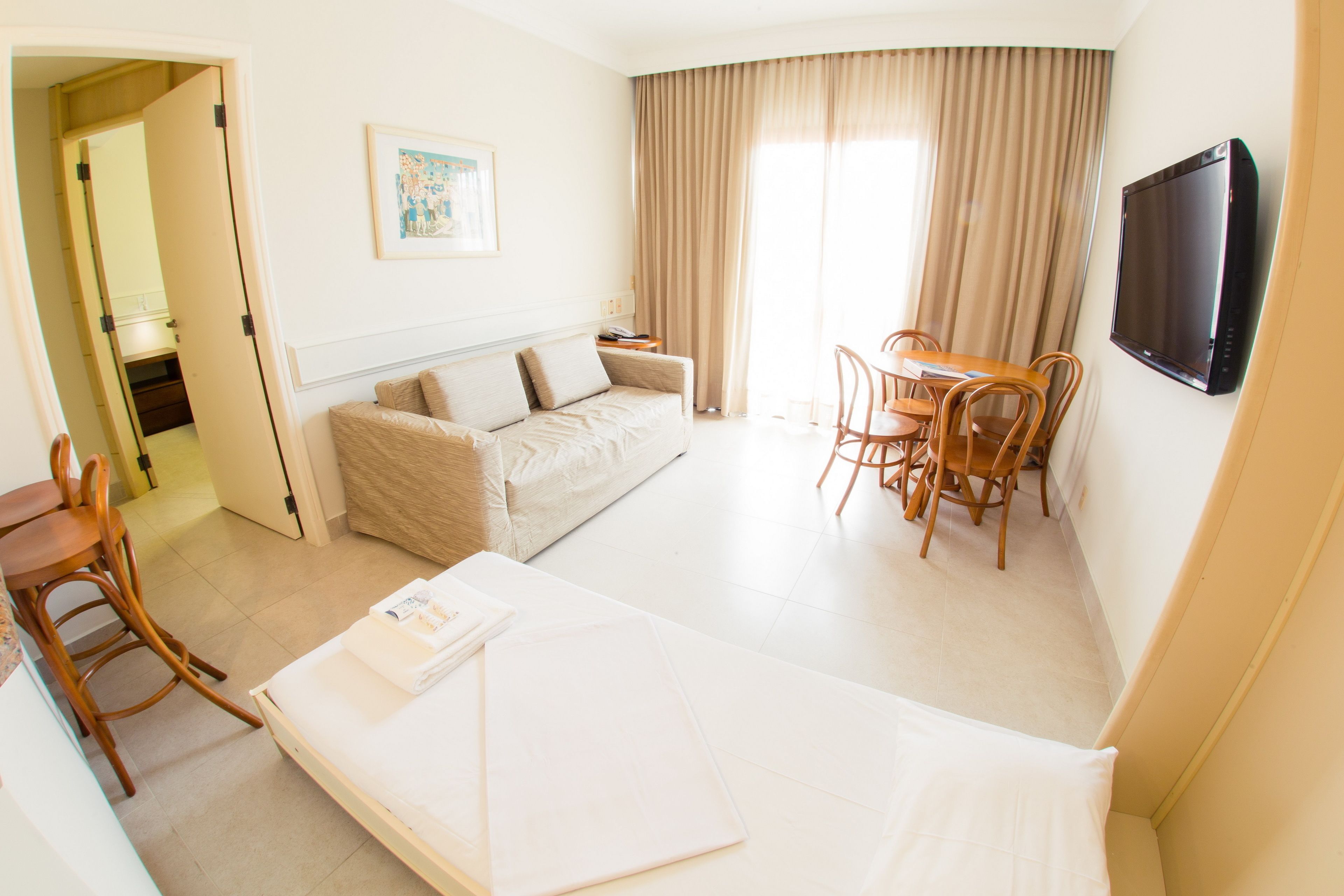 A hotel room with a bed, a sofa, a dining table with chairs, a TV, and a painting on the wall.