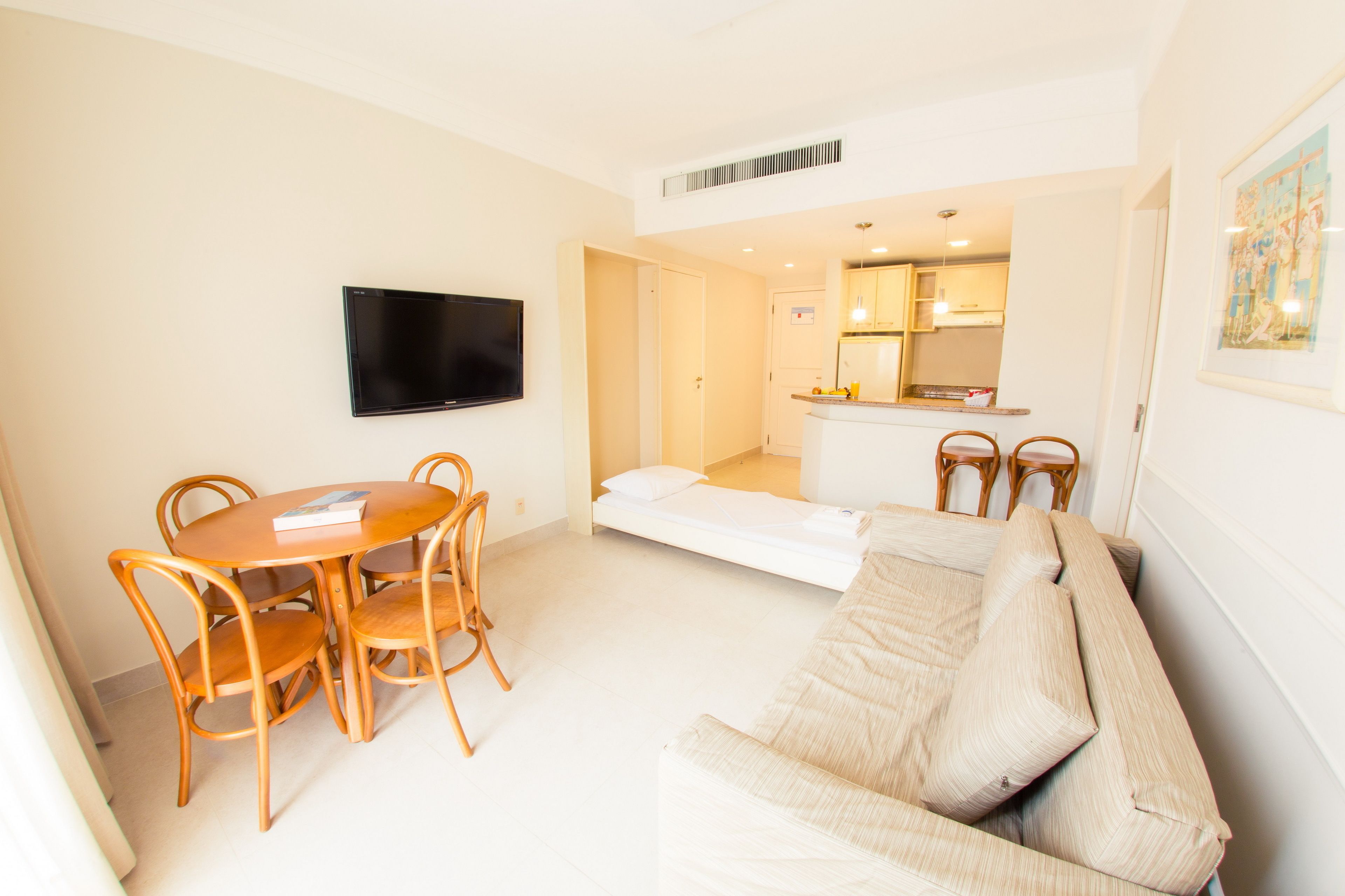 A modern living room with a sofa, dining table, and chairs, a flat-screen TV mounted on the wall, and a kitchenette in the background.