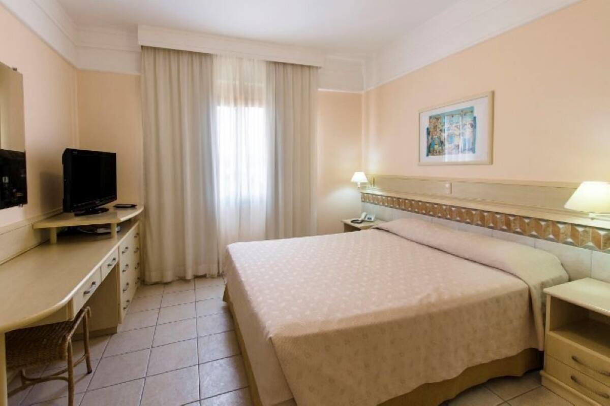 Ein Hotelzimmer mit Bett, Schreibtisch mit Fernseher, Nachttisch und einem Fenster mit Vorhängen.