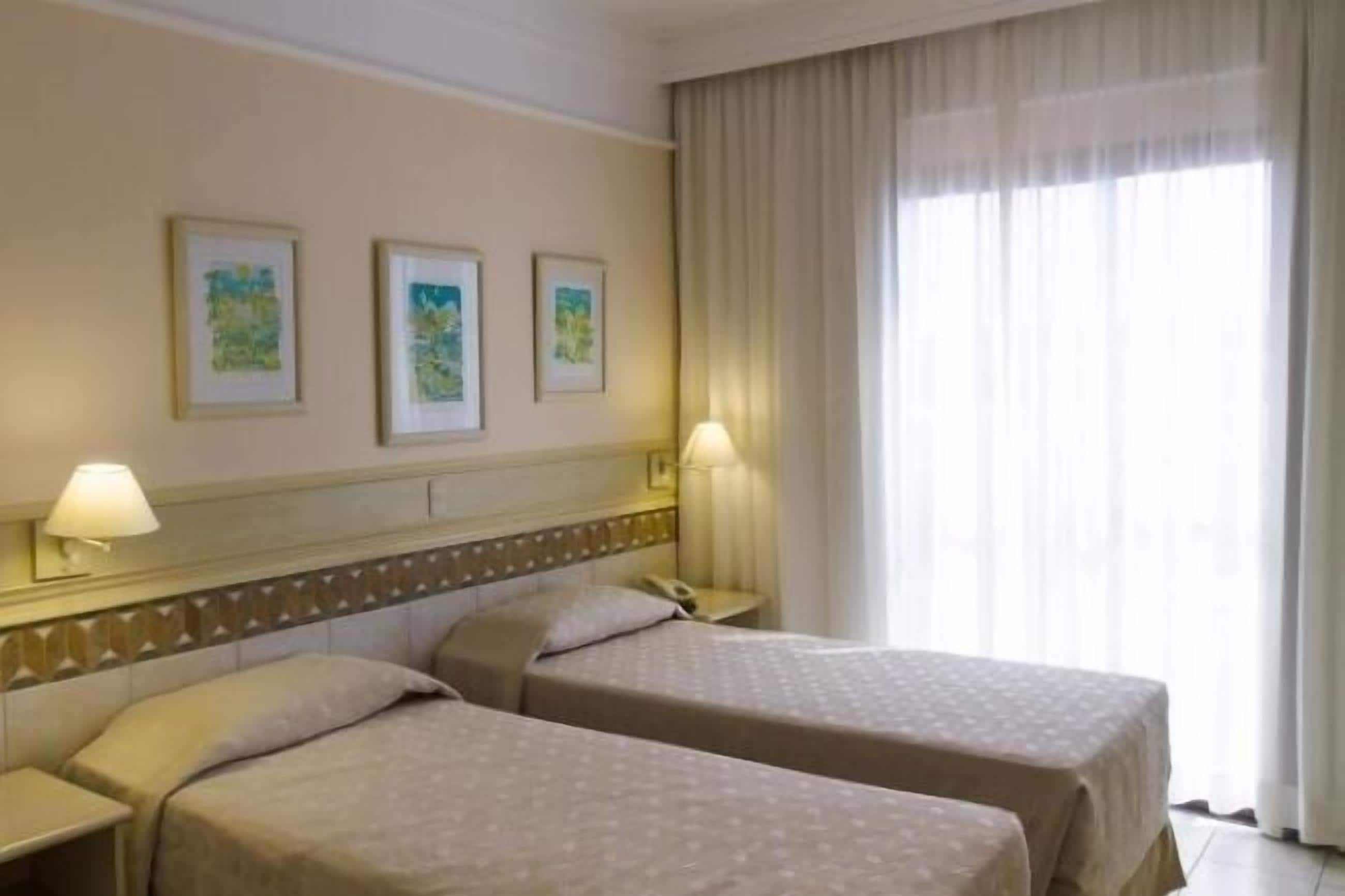 Une chambre d’hôtel avec deux lits, une tête de lit ornée d’un panneau décoratif, une table de chevet avec une lampe et une fenêtre avec des rideaux.