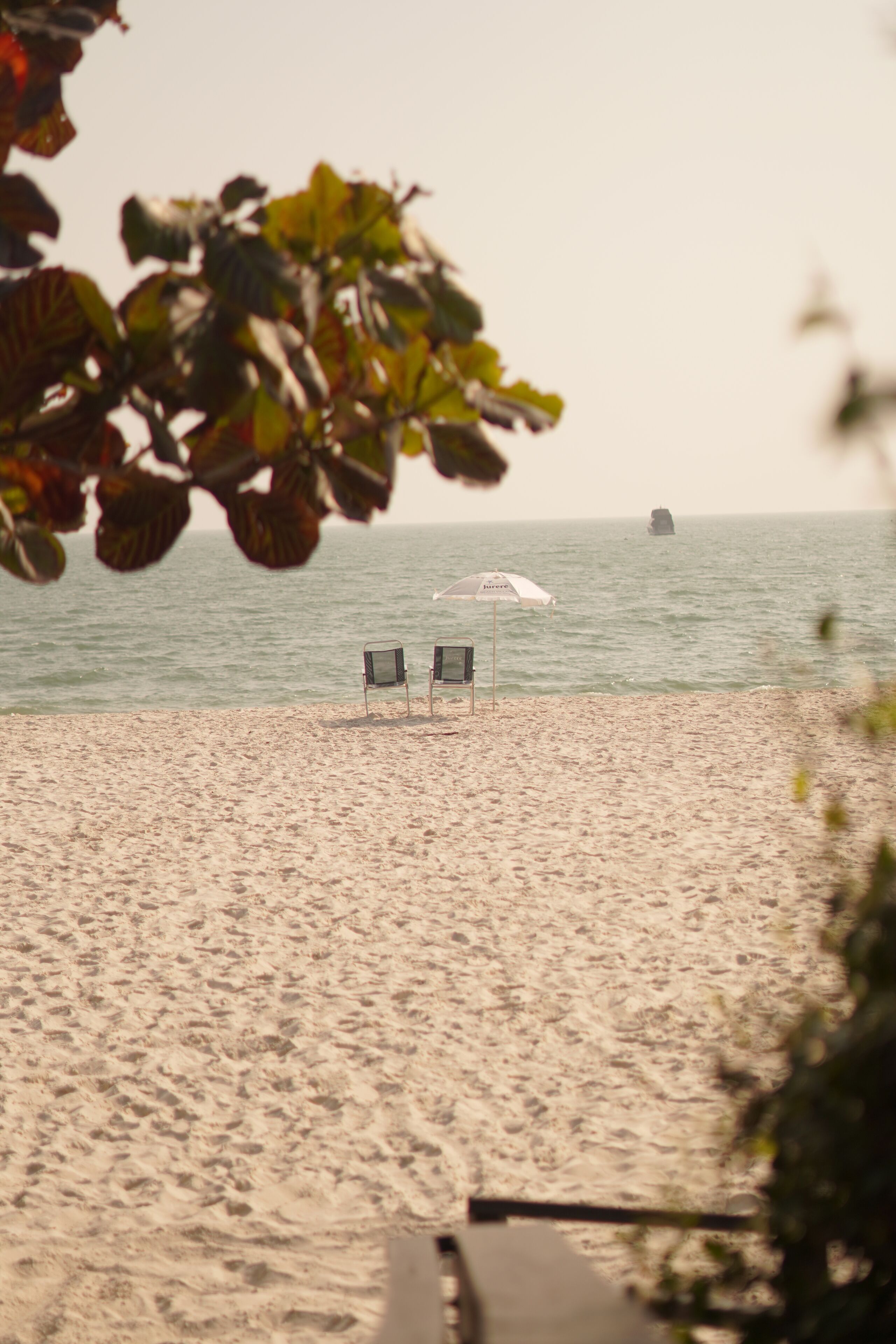 A beach with two chairs and an umbrella, a clear view of the ocean, and a distant rock formation.