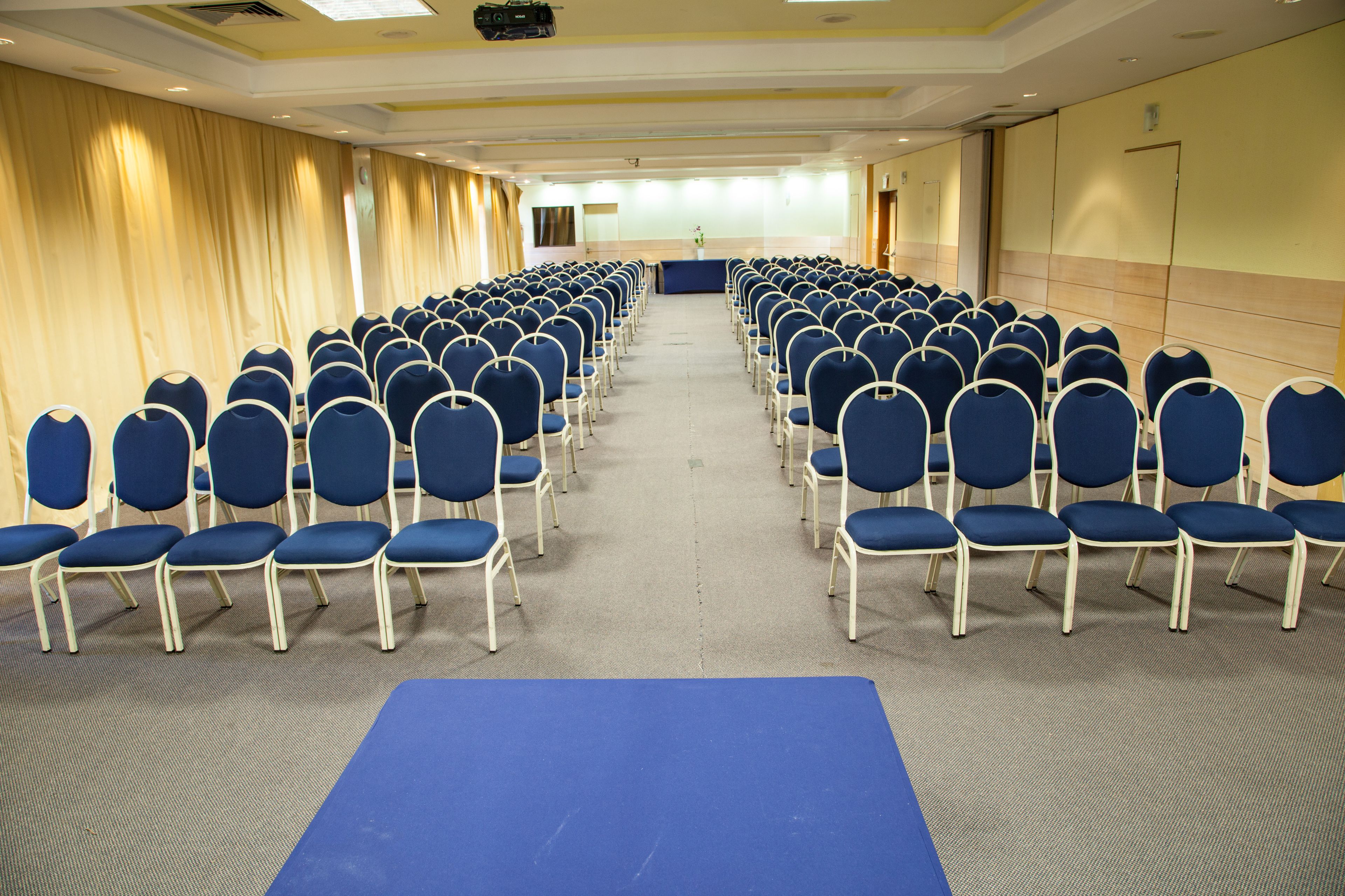 Un salón de conferencias con filas de sillas azules, un escenario con un atril y suelo alfombrado.