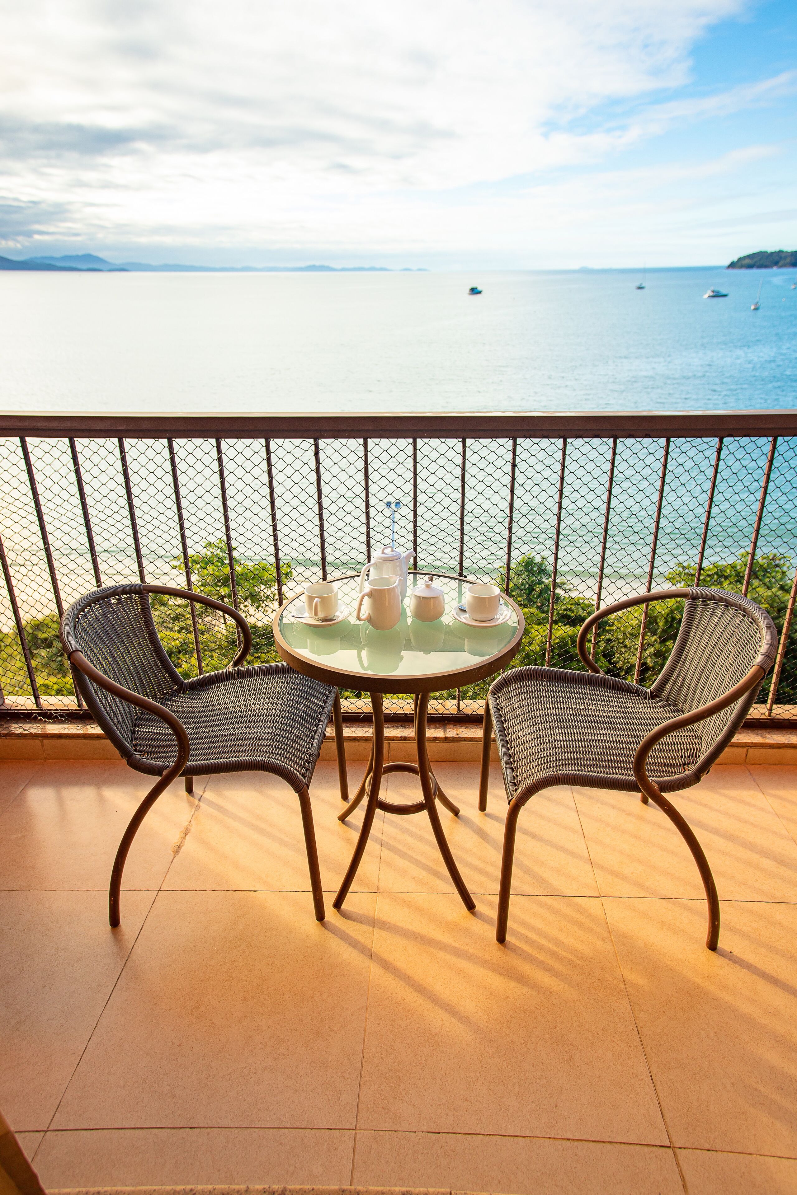 A balcony with two wicker chairs and a glass table, overlooking a calm sea with boats.