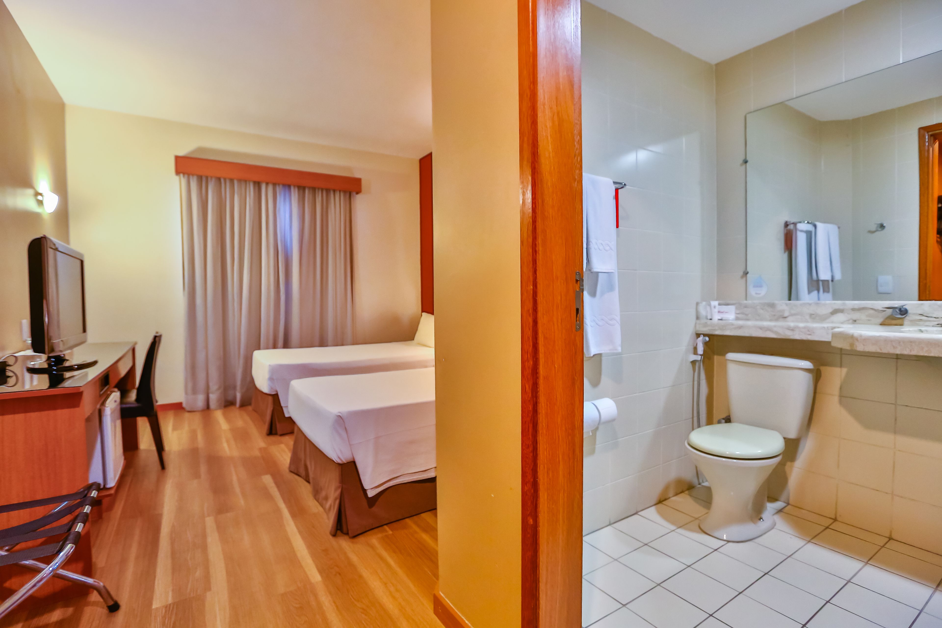 A hotel room with a wooden floor, a desk with a television, two beds, a bathroom with a toilet and sink, and a large mirror.