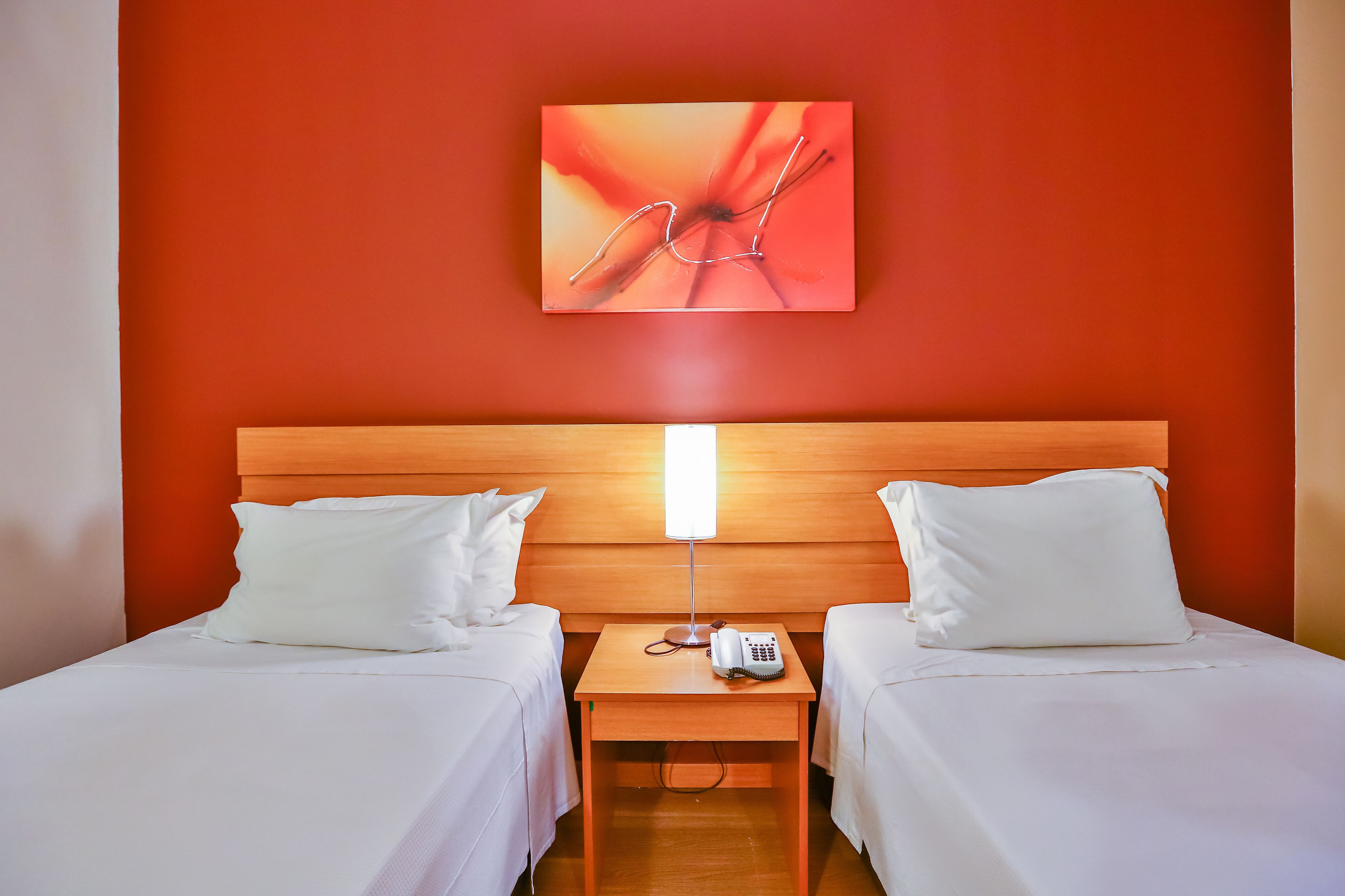 Quarto de hotel com duas camas, cabeceira de madeira, mesa de cabeceira com abajur e uma obra de arte pendurada na parede.