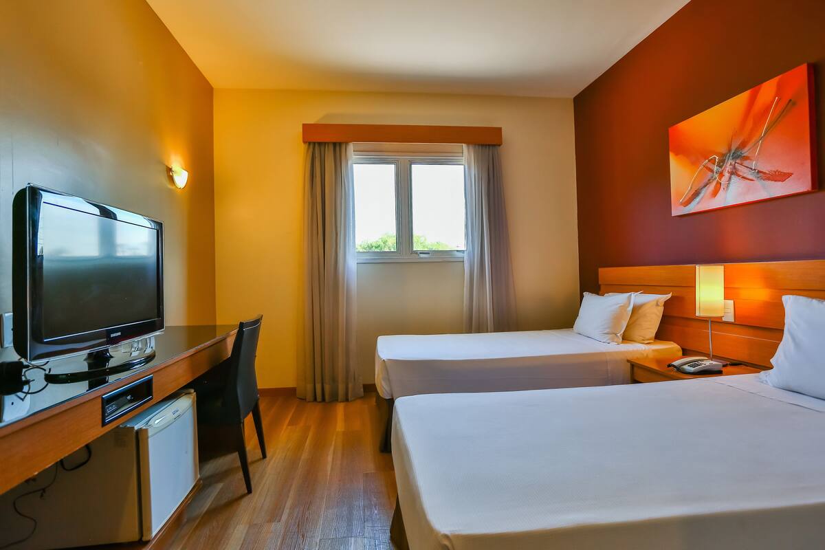 Habitación de hotel con dos camas, un escritorio con televisor, un refrigerador pequeño y una ventana con cortinas.