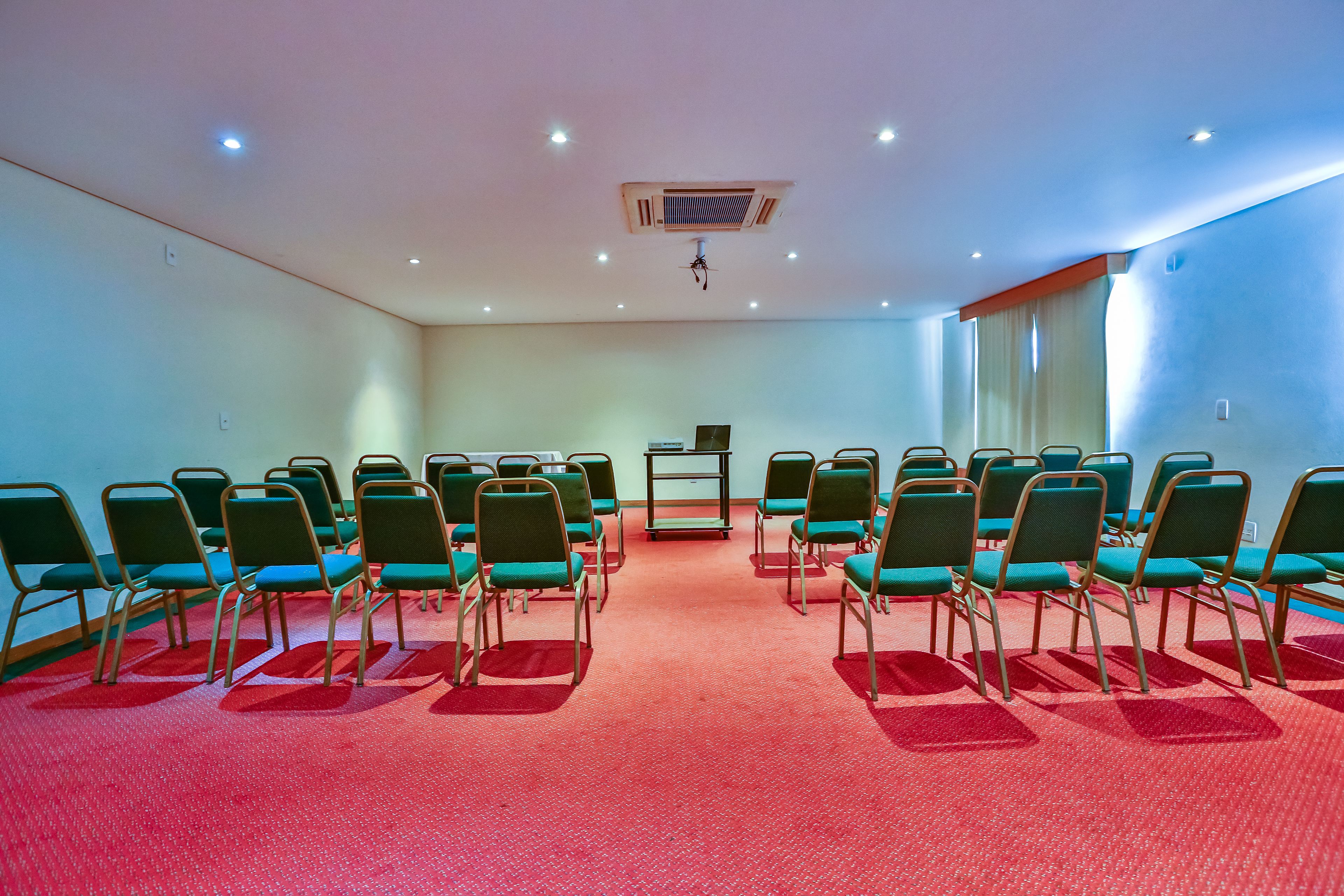 Een vergaderzaal met rijen groene stoelen, een rood tapijt en een plafondprojector.