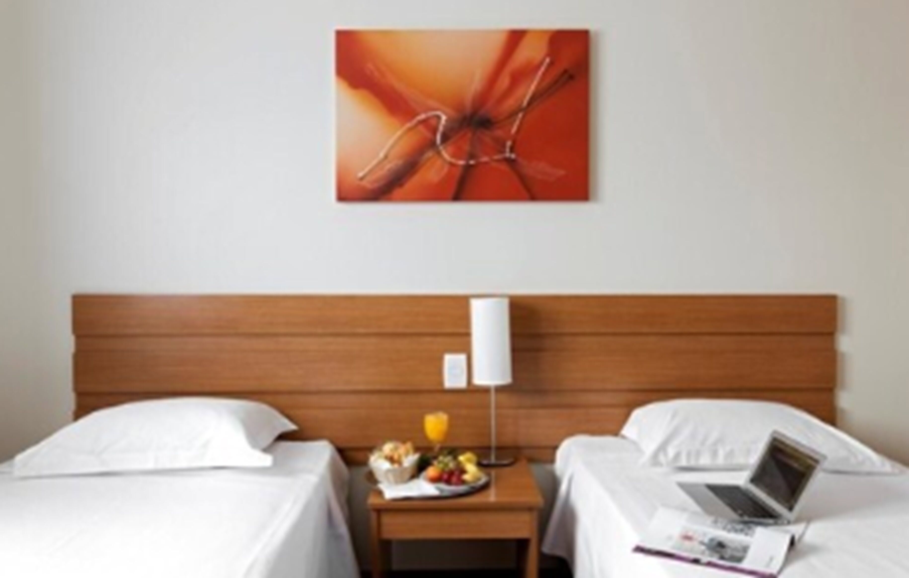 Quarto de hotel com cabeceira de madeira, duas camas com roupa de cama branca, uma mesinha de cabeceira com uma fruteira e um abajur, e um laptop sobre uma das camas.