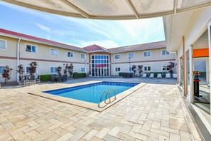 Une piscine avec une terrasse carrelée, entourée d’un bâtiment doté de nombreuses fenêtres et d’un toit rouge.