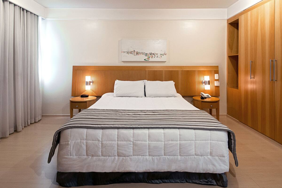 A hotel room with a large bed, wooden headboard, two bedside tables with lamps, a wardrobe, and a cityscape artwork on the wall.