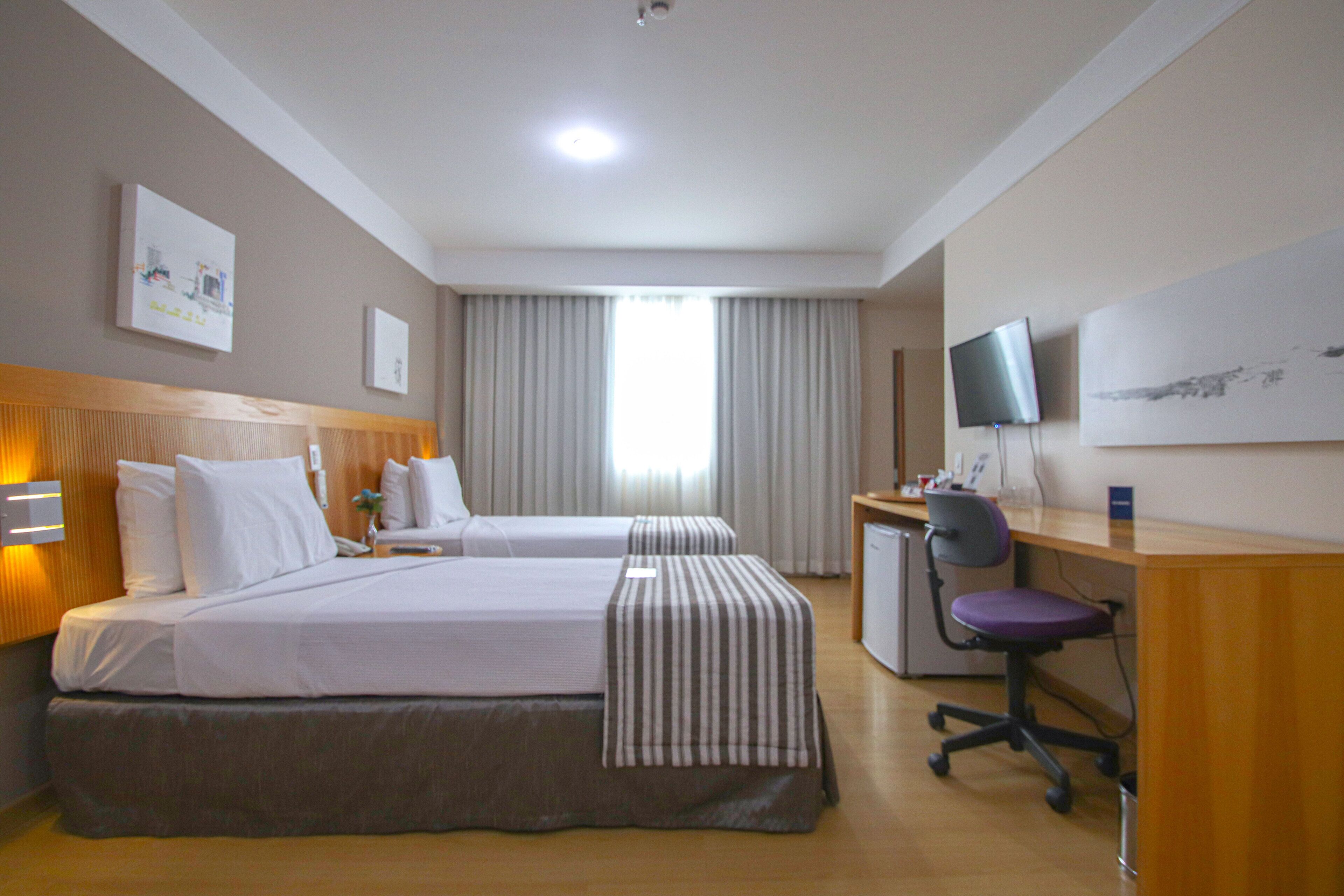 Habitación de hotel con dos camas, un escritorio, una silla, un televisor y una ventana con cortinas.