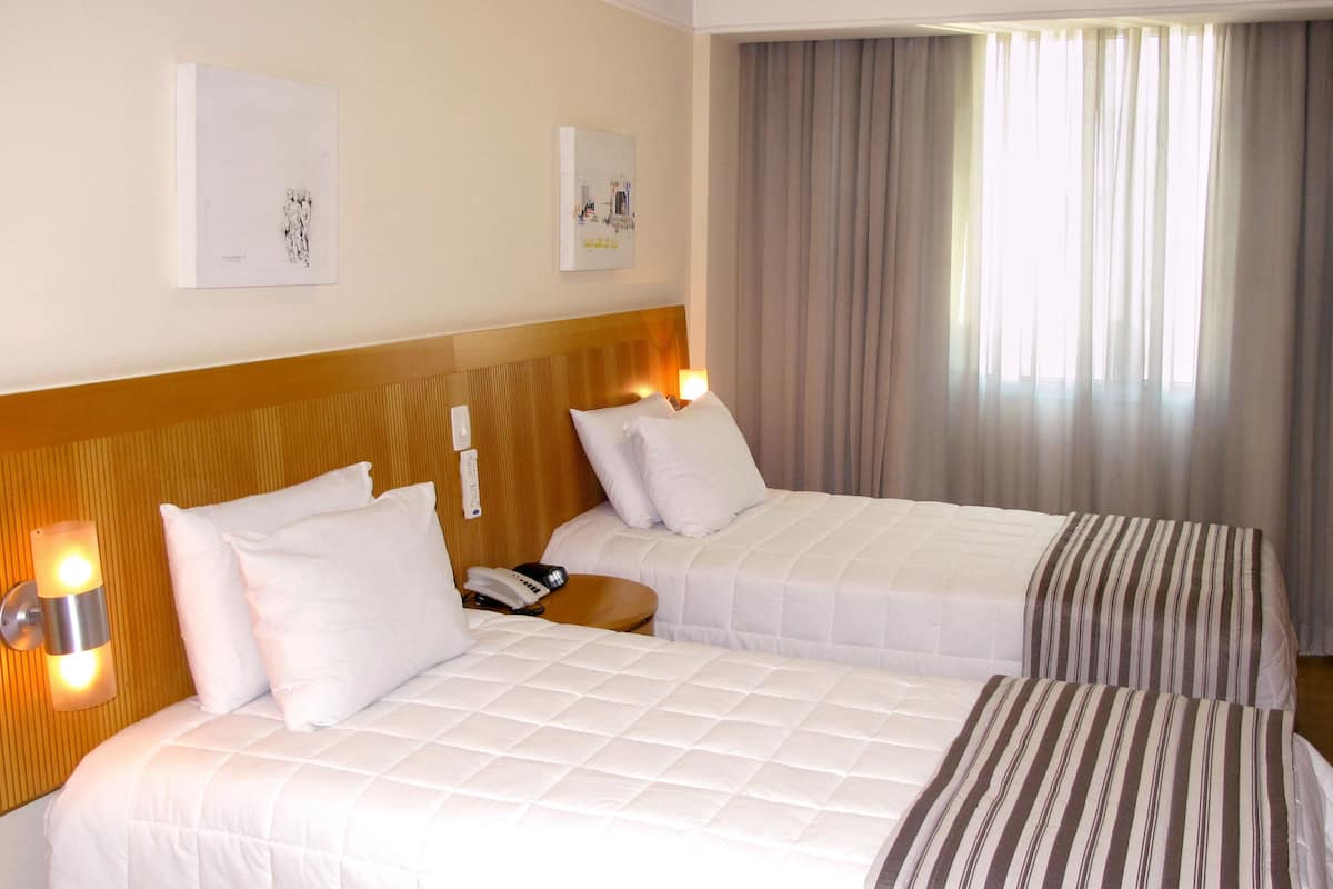 A hotel room with two beds, a wooden headboard, a bedside table with a lamp, and a window with curtains.