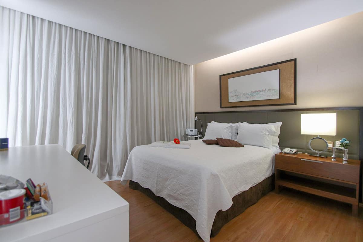 A hotel room with a large bed, a bedside table with a lamp, a desk with a chair, and a large window with curtains.