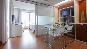 A modern hotel room with a glass dining table, white chairs, a flat-screen TV, and a large window with curtains.