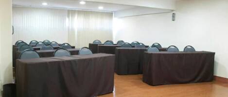 Una sala de conferencias con varias mesas y sillas, piso de madera y un ventanal con persianas.