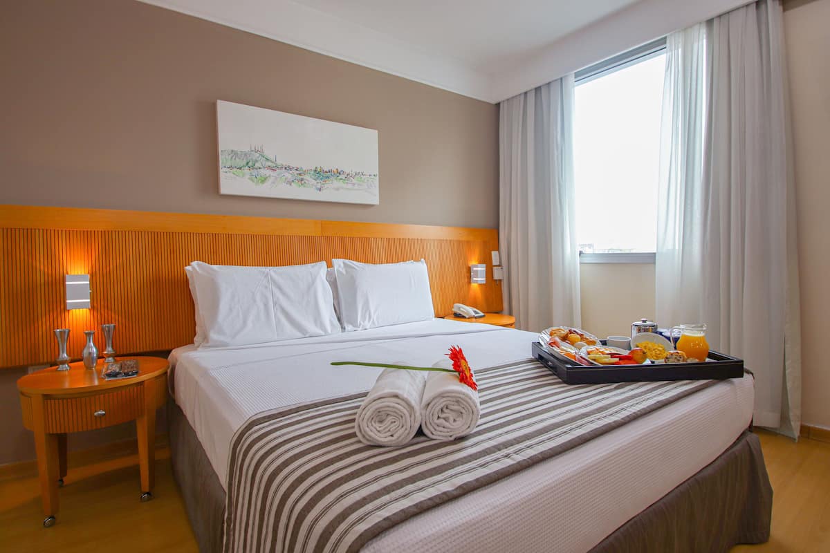 A hotel room with a bed, a bedside table with a lamp, a tray with breakfast items, and a painting on the wall.