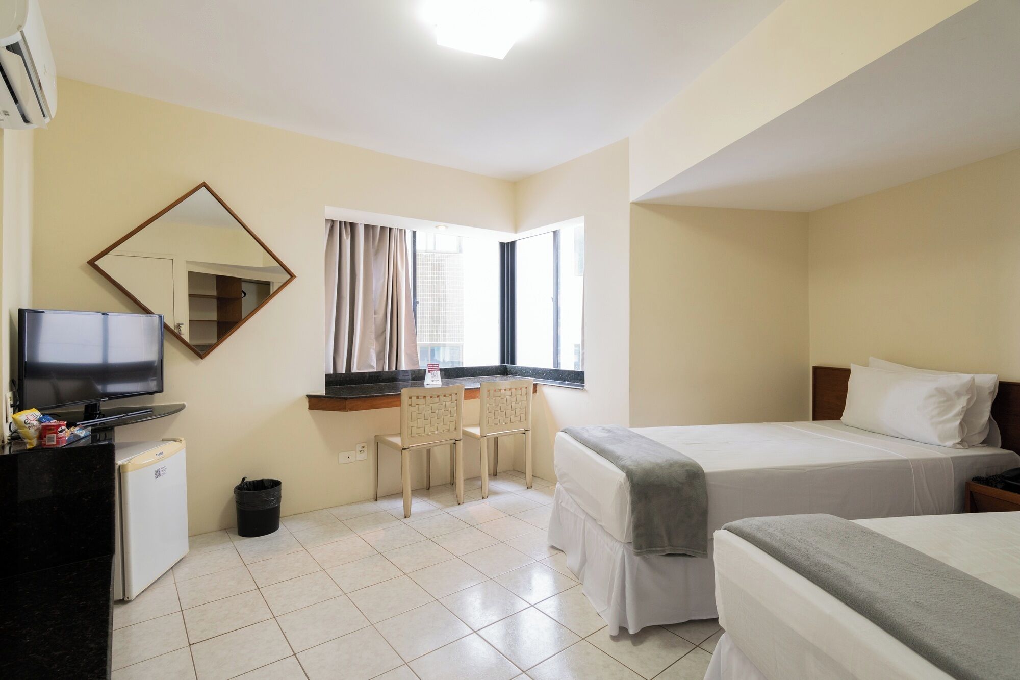 A hotel room with a bed, a desk with a television, a chair, a small trash bin, and a window with curtains.
