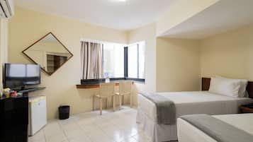 A hotel room with a bed, a desk with a television, a chair, a small trash bin, and a window with curtains.