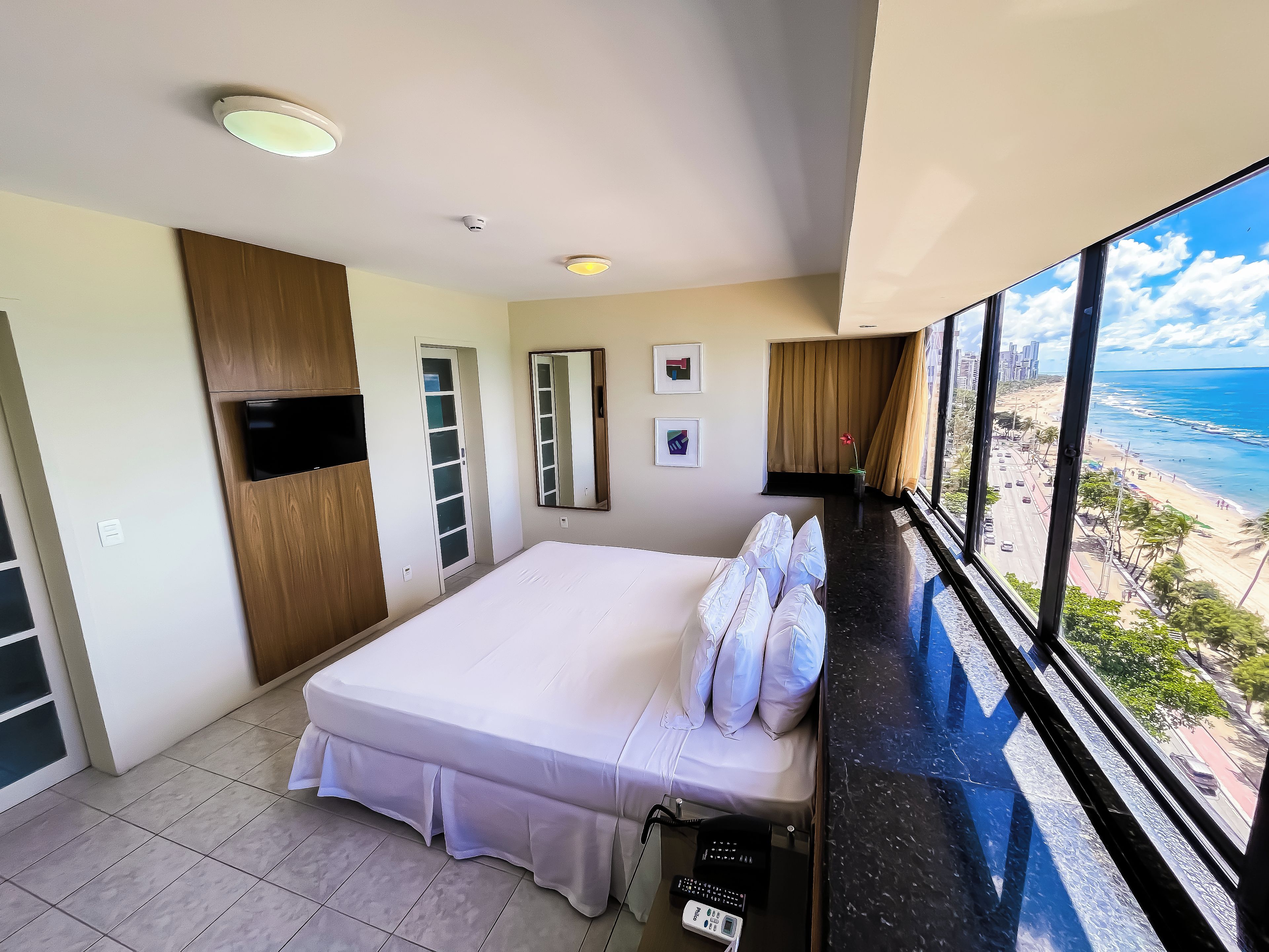 A hotel room with a bed, a TV, a wardrobe, a window with a beach view, and a balcony.