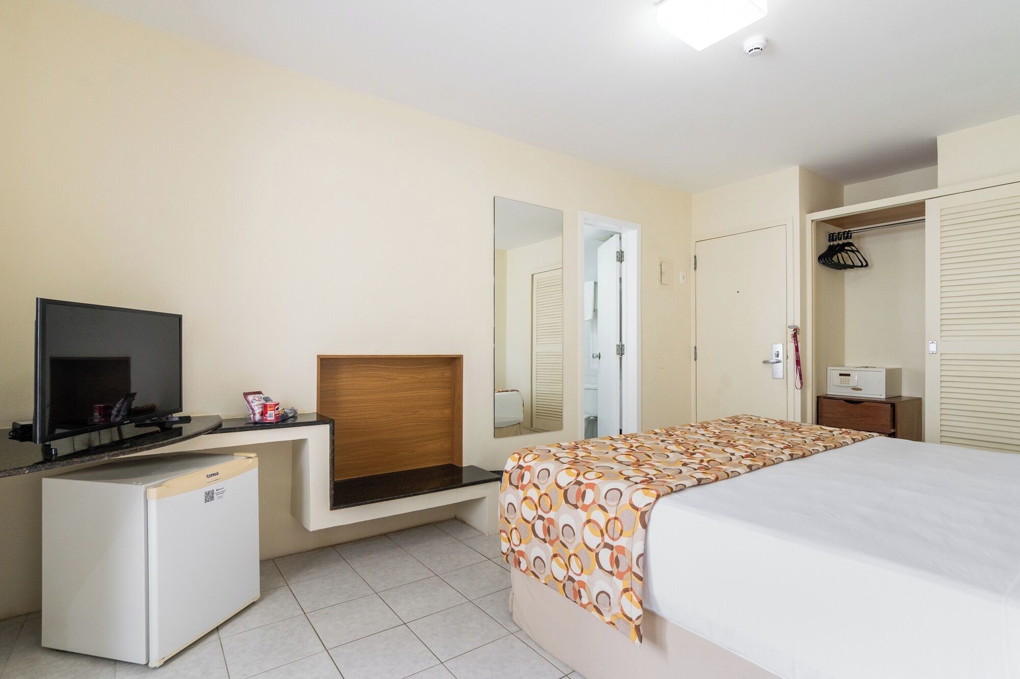 A hotel room with a bed, a TV, a minibar, a mirror, and a wardrobe.