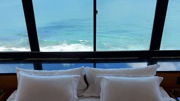 A bed with white linens and pillows, a view of the ocean through large windows. - Hotel Euro Suíte Recife By Nacional Inn (Recife)