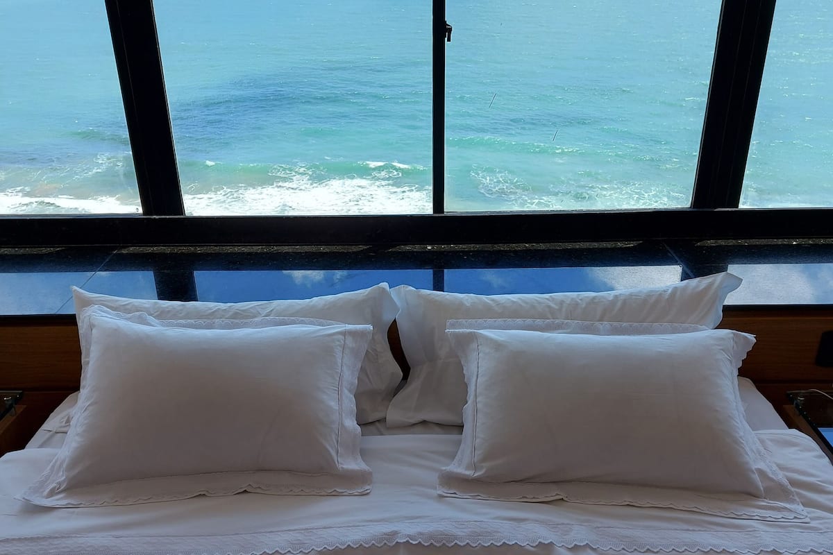 Una cama con sábanas y almohadas blancas, vista al océano a través de amplias ventanas.