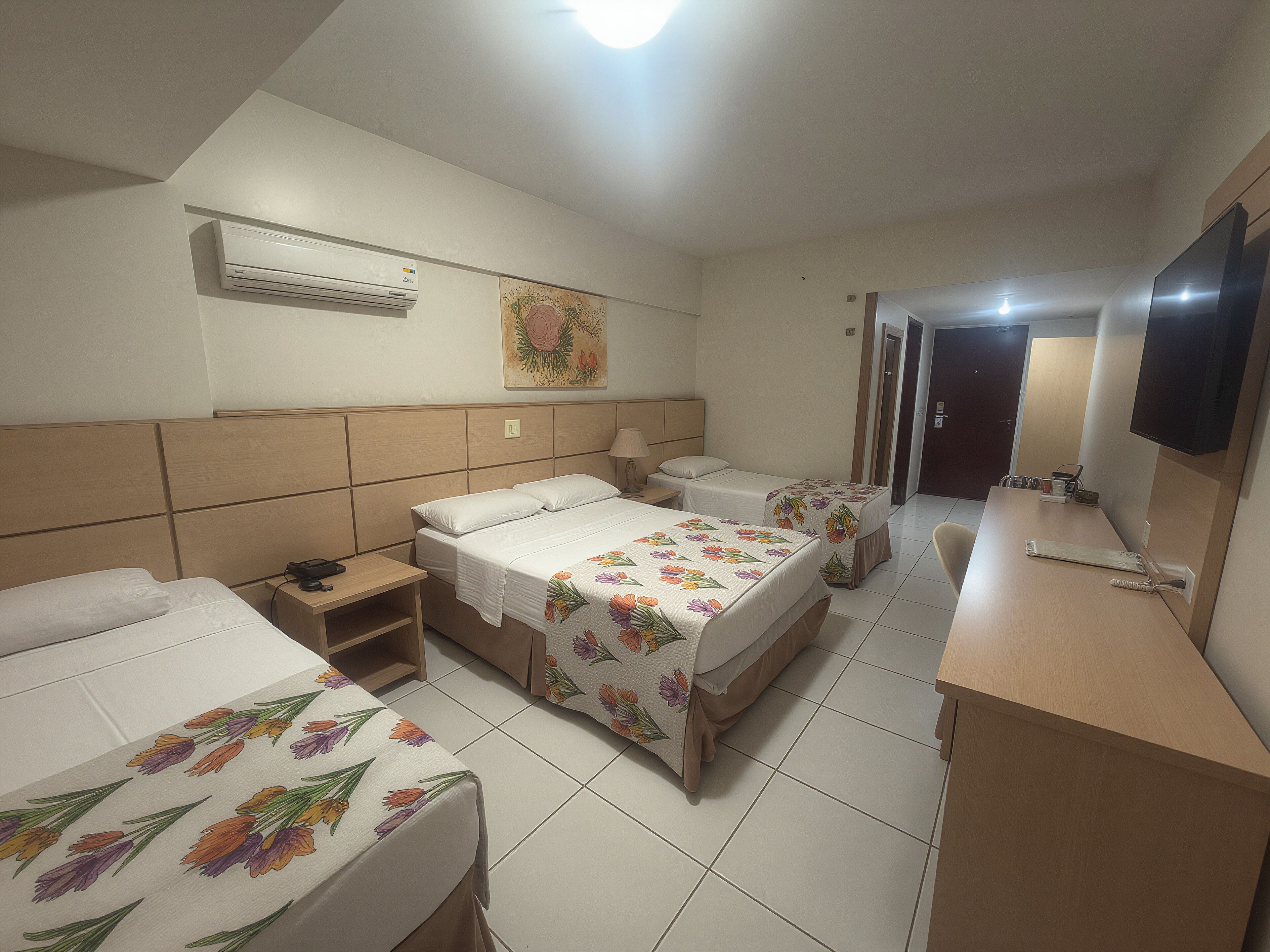 A hotel room with two beds, a desk, a TV, and an air conditioning unit.