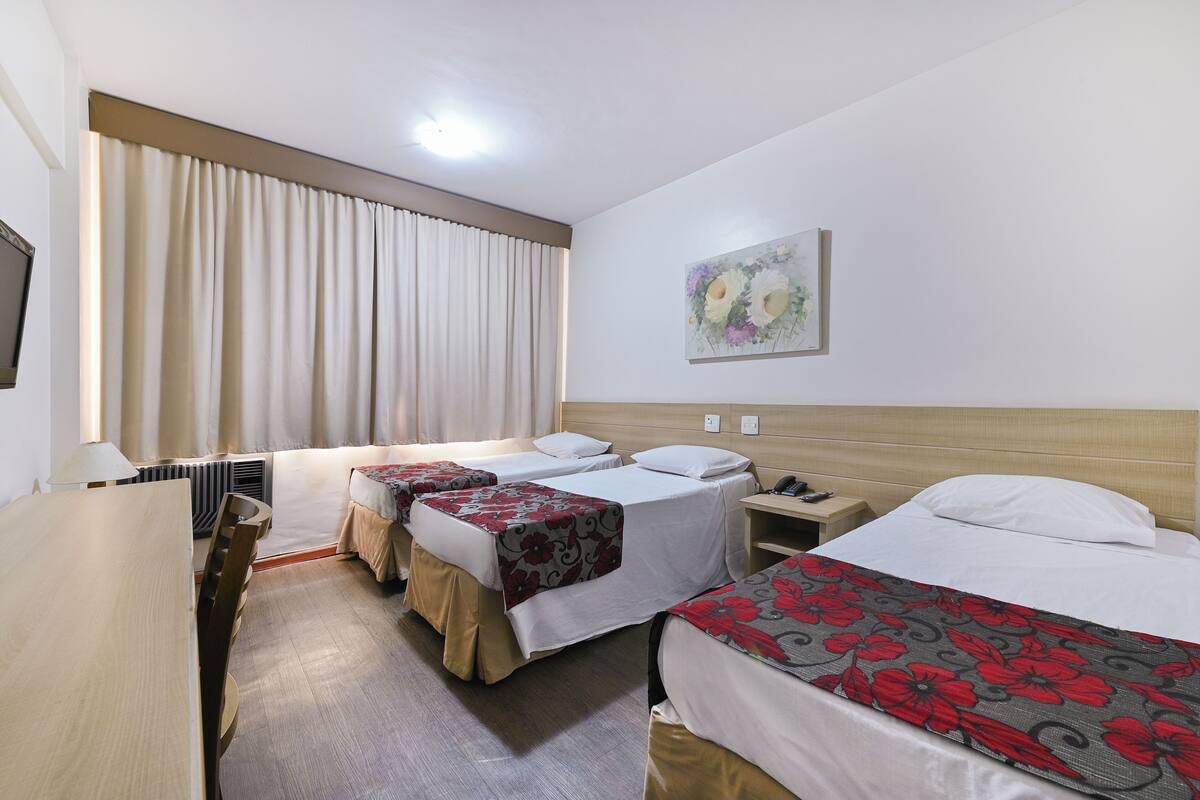 A hotel room with three single beds, a wooden desk, a chair, a bedside table, a lamp, and a painting on the wall.