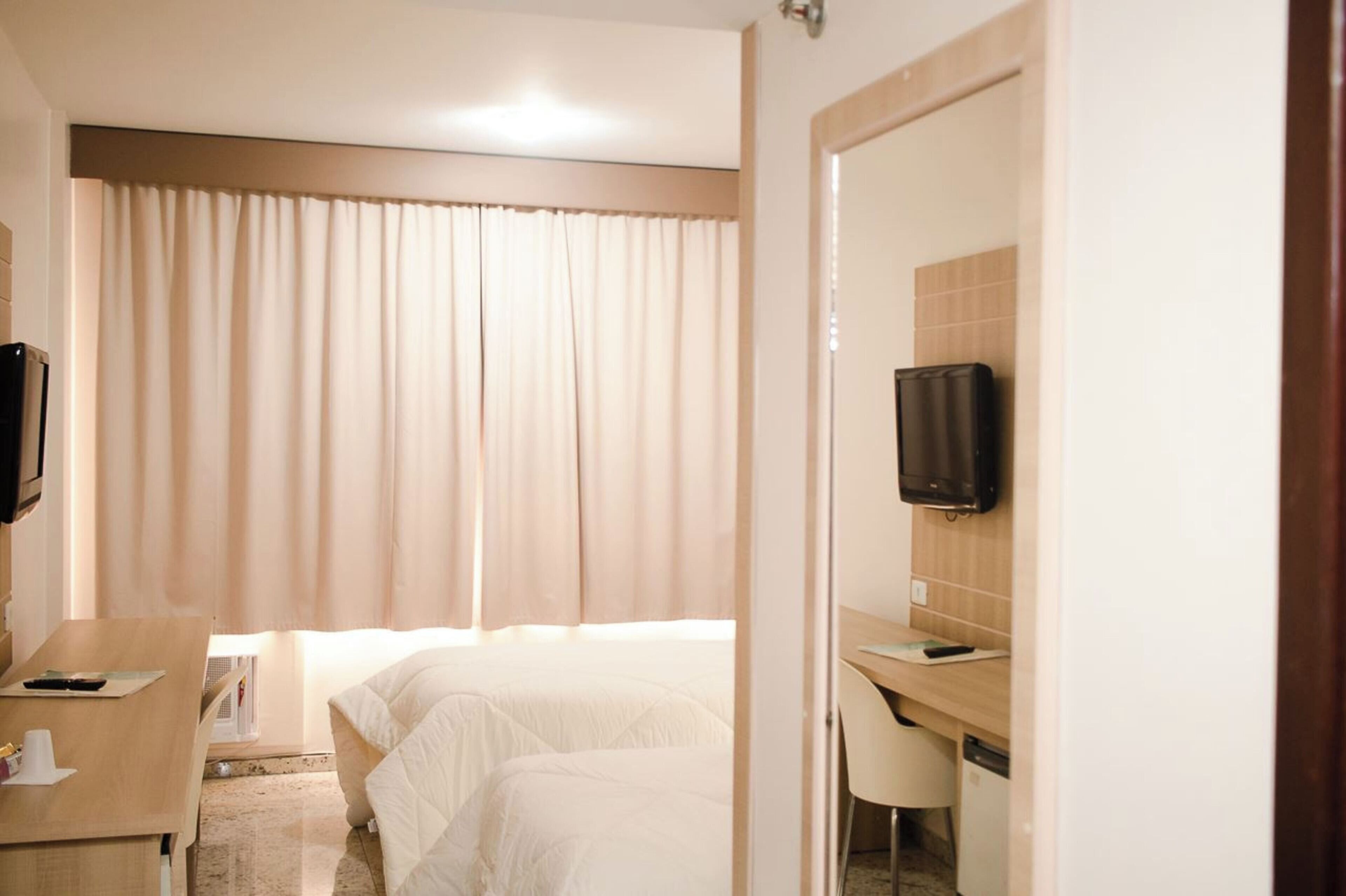 Una habitación de hotel con cama, escritorio, silla, televisión y ventana con cortinas.