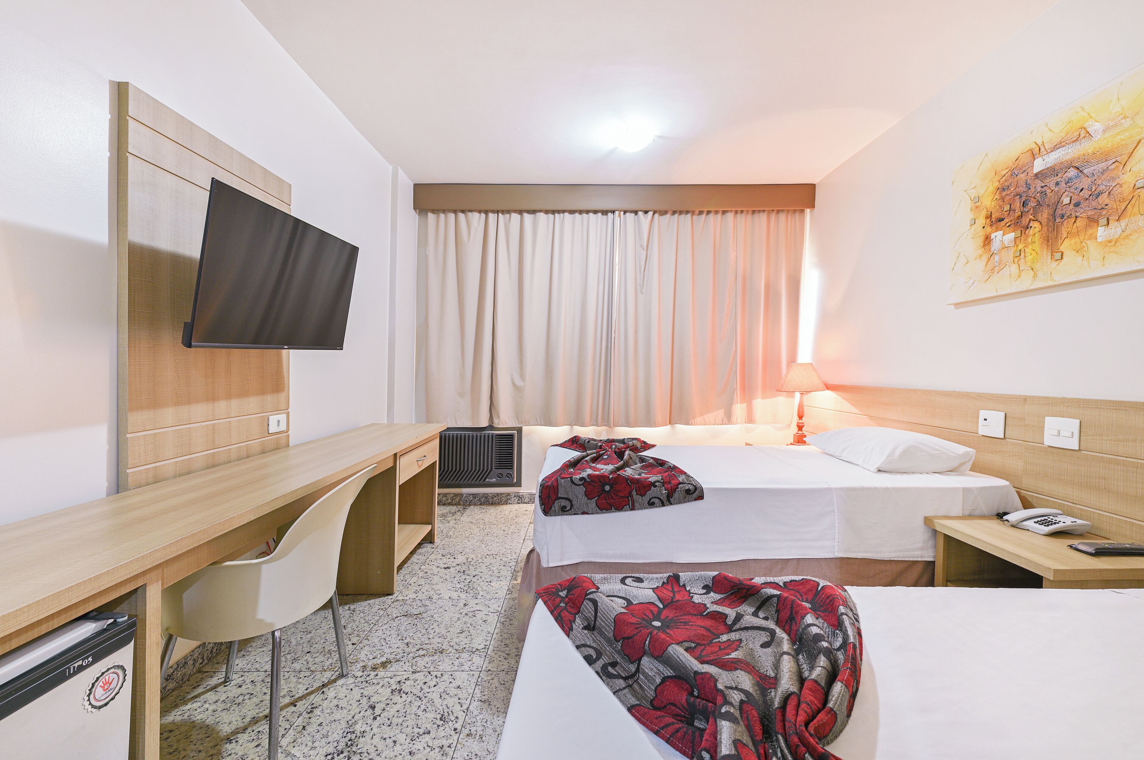 Quarto de hotel com duas camas, uma escrivaninha, uma cadeira, TV fixada na parede e um quadro na parede.