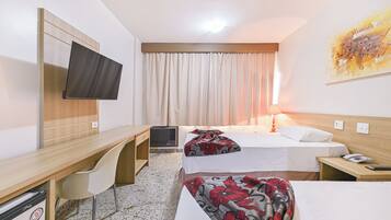 Quarto de hotel com duas camas, uma escrivaninha, uma cadeira, TV fixada na parede e um quadro na parede.