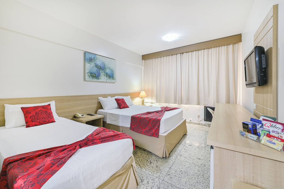A hotel room with two beds, a TV, a desk with a computer, and a window with curtains.