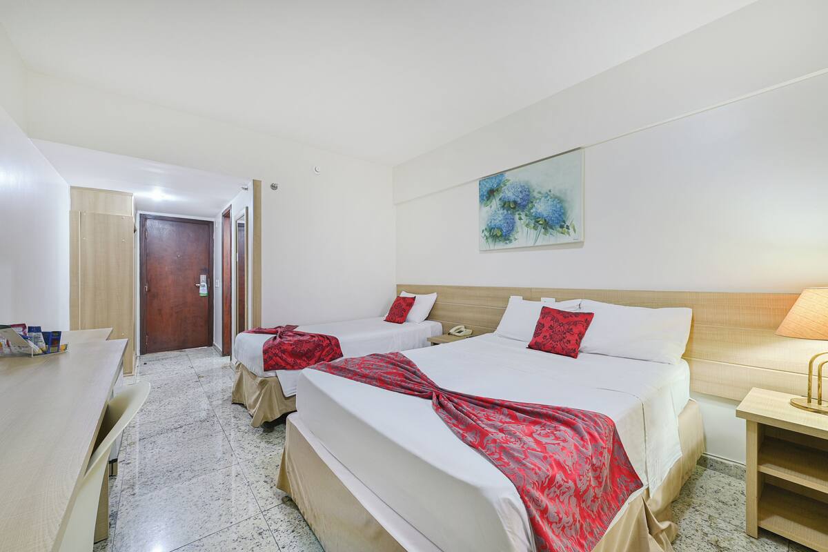 A hotel room with two beds, a desk, a chair, a painting on the wall, and a door leading to another room.