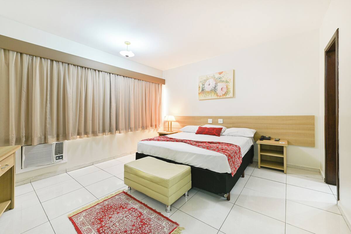 A bedroom with a bed, bedside tables, a nightstand, a window with curtains, a radiator, and a wall-mounted artwork.