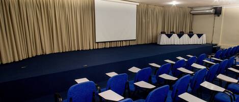 Une salle de conférence équipée d’une scène, de chaises bleues et d’un écran de projection.