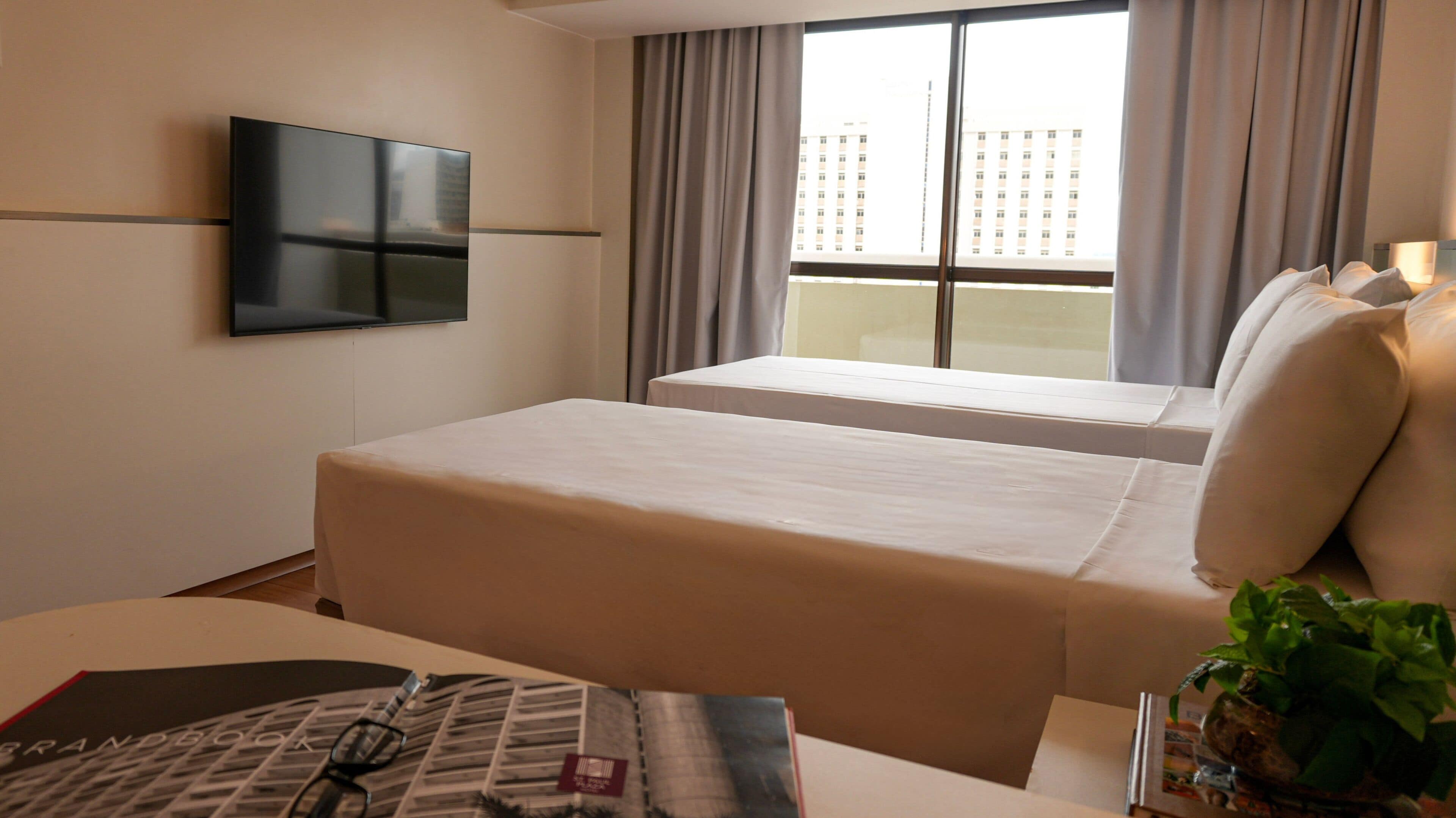 Habitación de hotel con cama, televisión en la pared, ventana con cortinas y planta sobre la mesita de noche.