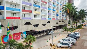 Um hotel de vários andares com varandas coloridas, área de estacionamento com carros e palmeiras.