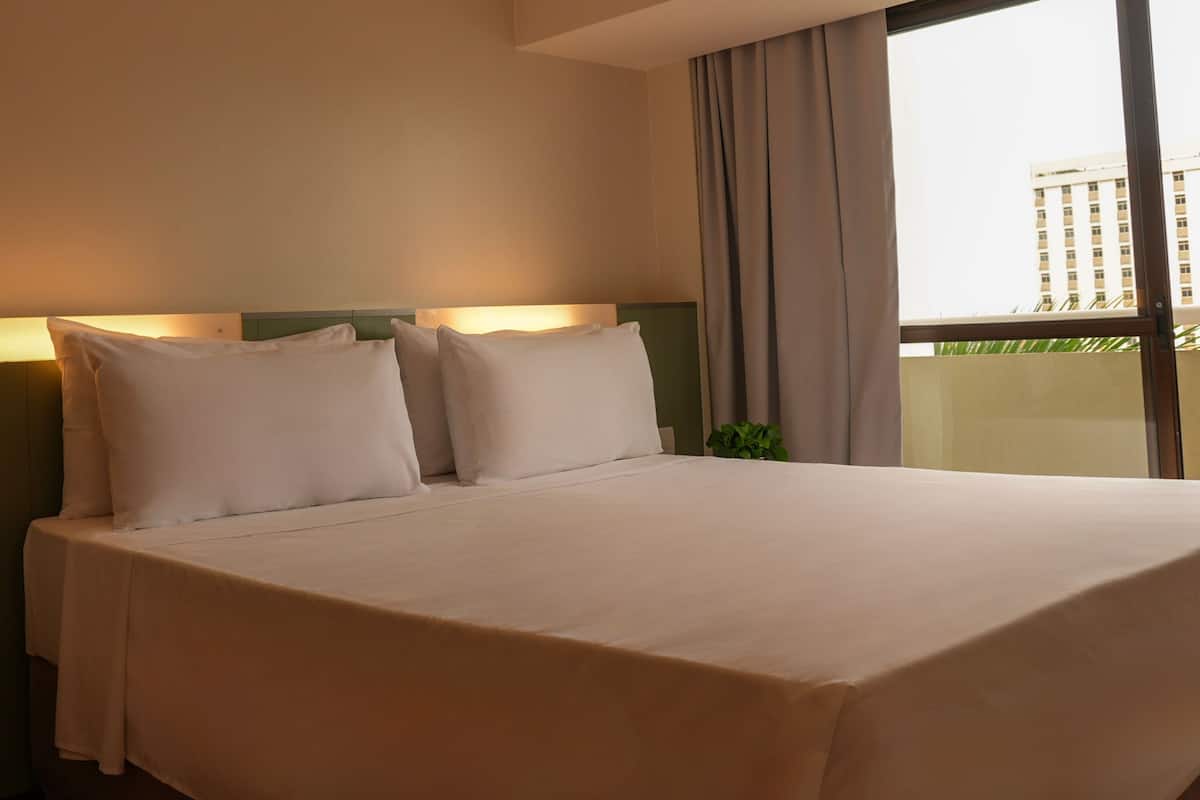 Ein Hotelzimmer mit einem großen Bett, zwei Kissen, einem Nachttisch mit Gläsern und Blick auf ein Gebäude durch das Fenster.