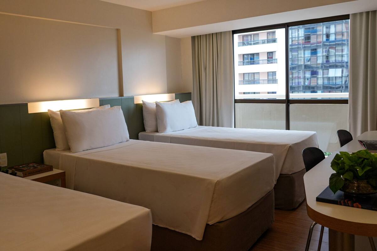 Ein Hotelzimmer mit zwei Betten, einem Schreibtisch und einem Fenster mit Blick auf Gebäude.