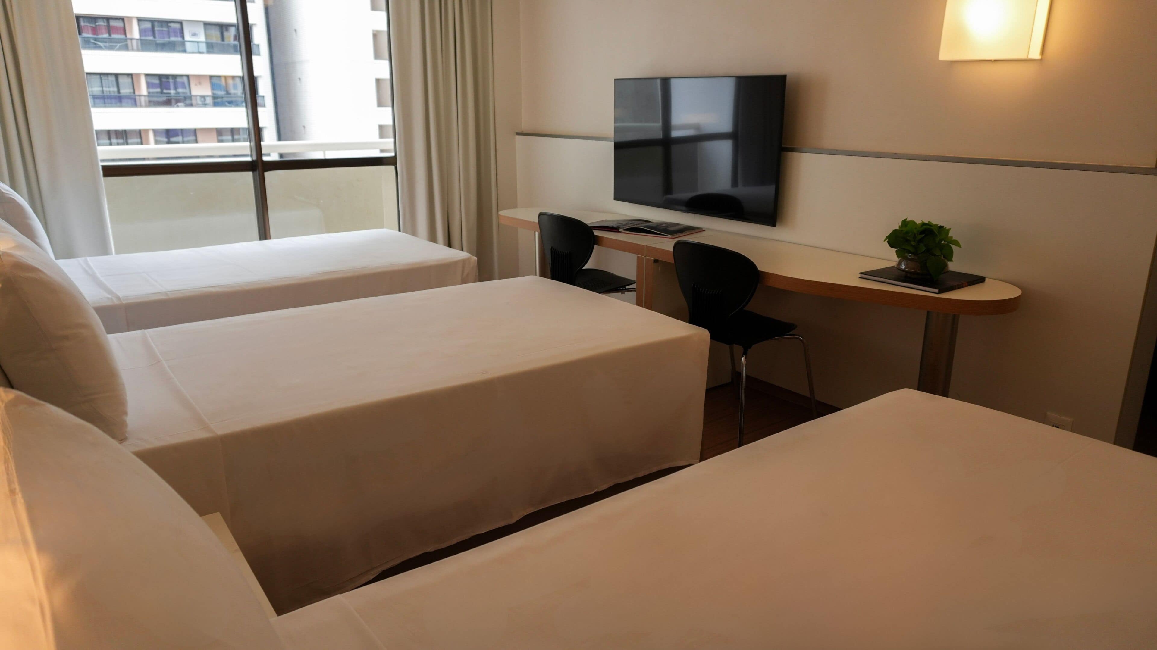 A hotel room with two beds, a desk, a chair, a TV, and a window with a view of buildings.