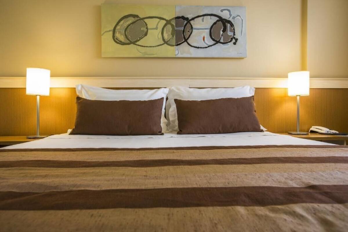Quarto de hotel com uma cama grande, duas luminárias laterais e arte abstrata na parede.