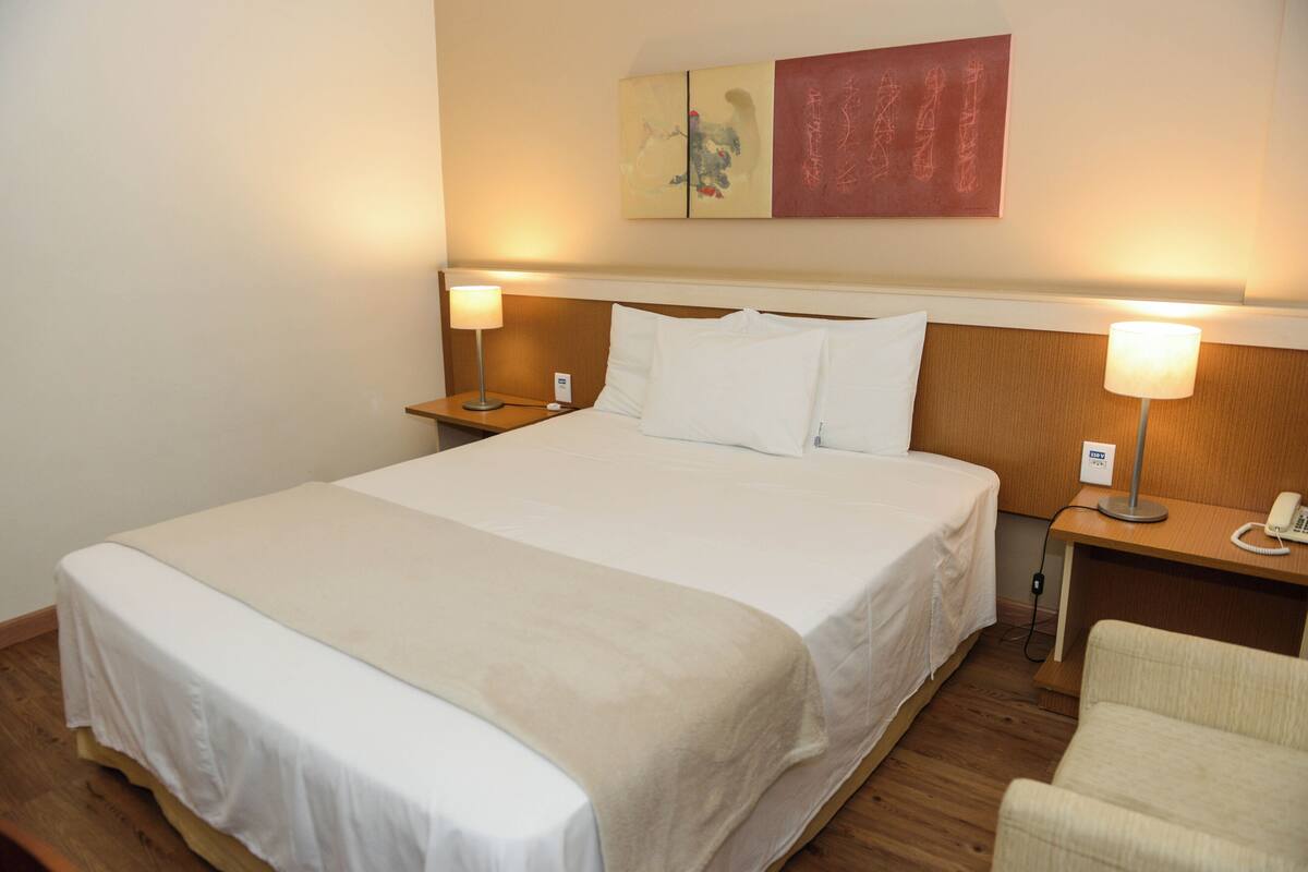 Quarto de hotel com uma cama grande, duas mesas de cabeceira com luminárias, uma cadeira e uma obra de arte na parede.